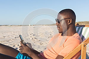 Happy african american man in sunglasses sitting in deckchair using smartphone on sunny beach