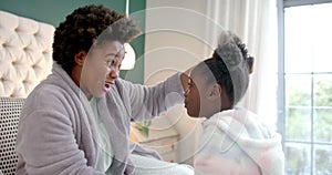 Happy african american mother combing daughter hair sitting on bed in bedroom, slow motion