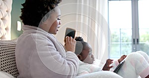 Happy african american mother combing hair of daughter using tablet in bedroom, slow motion