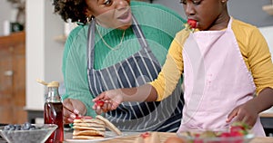 Happy african american mother and daughter decorating pancakes with fruits in kitchen, slow motion