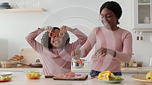 Happy african american mother and daughter taking slices of tomato and making vegetable glasses, smiling to camera