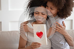 Happy african american mother hugging daughter, holding greeting card.