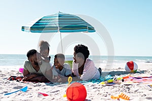 Happy african american parents and children talking while lying on towels at sandy beach under sky
