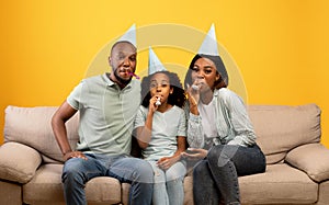 Happy african american parents and girl celebrating birthday and blowing at party horns over yellow background