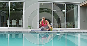 Happy african american plus size woman practicing yoga, using smartphone next to swimming pool