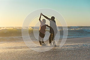 Happy african american retired senior couple dancing on shore at beach against sky during sunset