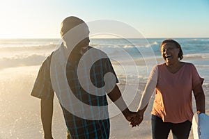 Happy african american senior couple holding hands walking together at beach during sunset