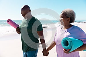 Happy african american senior couple with yoga mats holding hands and walking at beach, copy space