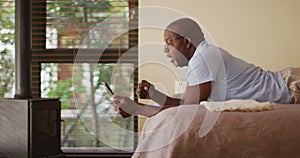 Happy african american senior man lying on bed watching tablet and cheering