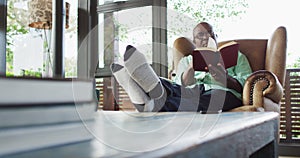 Happy african american senior man relaxing in armchair with feet up, reading a book