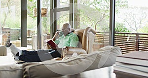 Happy african american senior man relaxing in armchair, feet up, reading book and drinking coffee