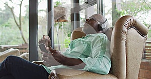 Happy african american senior man relaxing in armchair, listening using headphones and smartphone