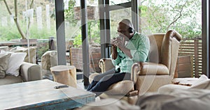 Happy african american senior man relaxing in armchair, wearing headphones and drinking coffee
