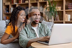 Happy african american spouses sitting by couch with laptop, looking for new furniture online, websurfing, copy space