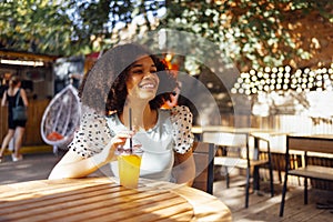 Happy african american teenage girl drinking lemonade or cocktail in an open air cafe. Smiling darkskin female teenager sits at