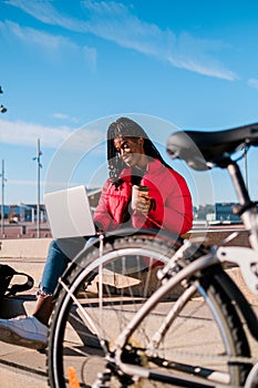 happy african-american woman with bike sitting on street stairs and working with a laptop