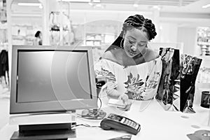 Happy african american woman customer with colored shopping bags paying by credit card near cash teminal with pos