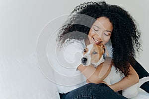 Happy African American woman expresses love to dog, embraces pet, dressed in casual wear, sit on comfortable bed in bedroom,