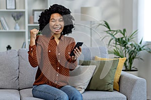 Happy African American woman holding phone while sitting on sofa at home and looking happy at camera showing victory