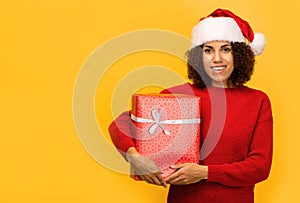 Happy African American woman holds in hands christmas gift Female in santa claus hat on orange background, smiling. Xmas