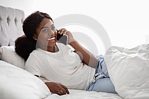 Happy african american woman laying in bed, having phone call