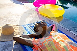 Happy african american woman lying on towel and sunbathing next to swimming pool in garden