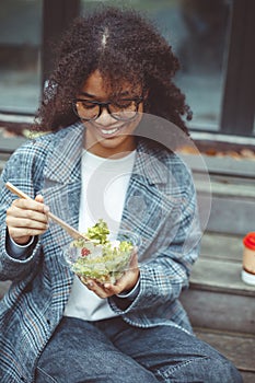 Happy african american woman office worker eating salad outdoors while sitting on bench in park