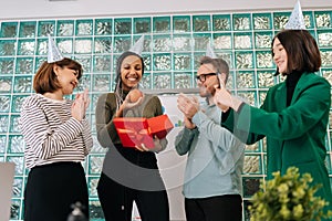 Happy African-American woman opening present given by multiethnic coworkers in modern office to applause of colleagues