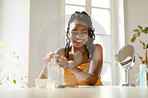 Happy african american woman pouring moisturising body lotion from bottle on hand, using new skincare product after bath