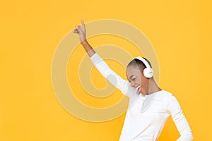 Happy African American woman raising hand up while listening to music in wireless headphones on yellow  studio