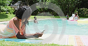 Happy african american woman relaxing at pool with family, using smartphone