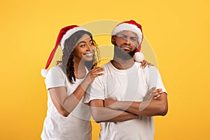 Happy african american woman in santa hat comforting her upset husband over yellow background