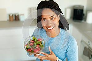 Happy african american woman in sportswear eating healthy salad after domestic workout, sitting in kitchen and smiling