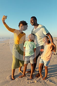 Happy african american woman taking selfie with family from smartphone at beach against blue sky