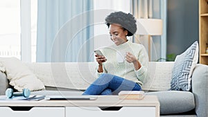 Happy African American woman texting on a phone while relaxing on a sofa and drinking coffee at home. Smiling black