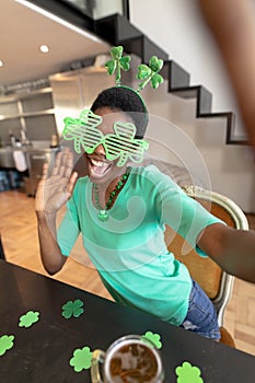 Happy african american woman wearing shamrock novelty glasses while taking selfie at home