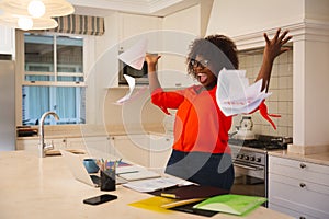 Happy african american woman working in kitchen with laptop, celebrating success, throwing papers