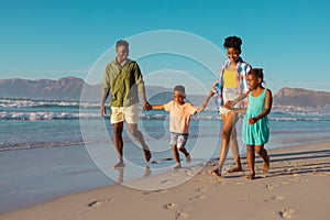 Happy african american young parents holding children's hands and walking at beach against clear sky