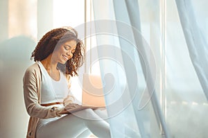 Happy african american young woman freelancer on windowsill with laptop on laps, working from home