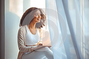 Happy african american young woman freelancer on windowsill with laptop on laps, working from home