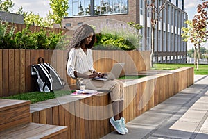 Happy African black girl student studying online using laptop sitting outdoors.