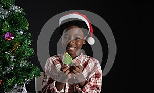 Happy African boy smile and holding Christmas tree shape cookie in hand near little Christmas tree on black background