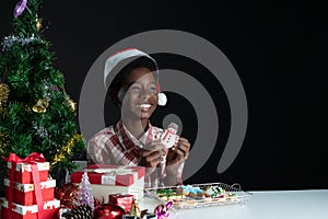 Happy African boy smile and holding snowman shape cookie in hand near little Christmas tree on black background