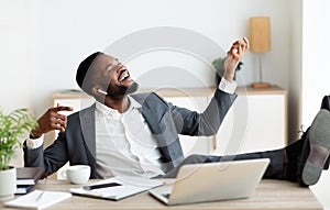 Happy african businessman listening to music with legs on table