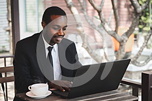 Happy african businessman in a suit use business laptop at coffee shop. Smiling black male executive typing business email on