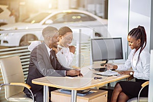 Happy african couple with dealer buying car in auto show or salon