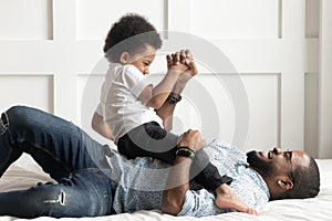 Happy african dad laughing playing with little son on bed