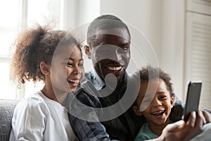 Happy african dad laughing using smartphone with kids at home