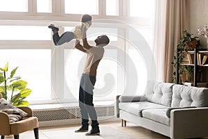 Happy african dad lifting little toddler son playing at home