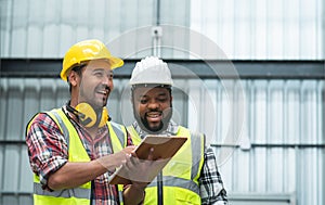 Happy African engineer and Asian foreman in safety hat working together at warehouse while listing on clipboard to discuss,